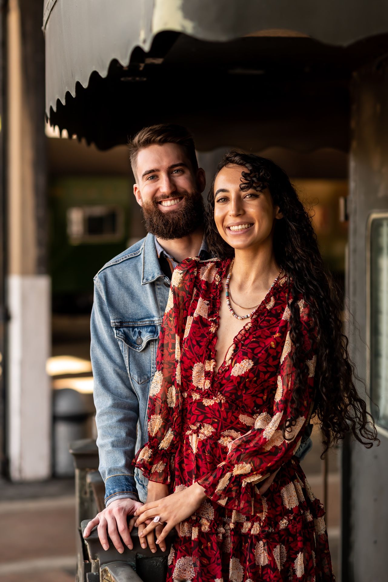 A couple in casual attire poses together under an outdoor overhang, with one wearing denim and the other in a red patterned dress.