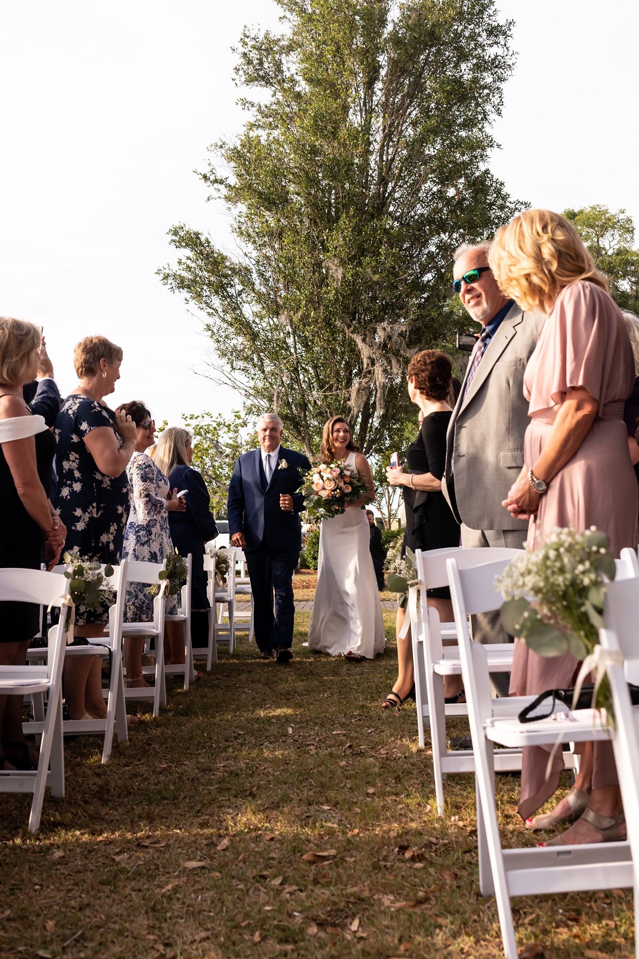 An outdoor wedding ceremony setup with white folding chairs and floral decorations lining a grass aisle under tall trees.