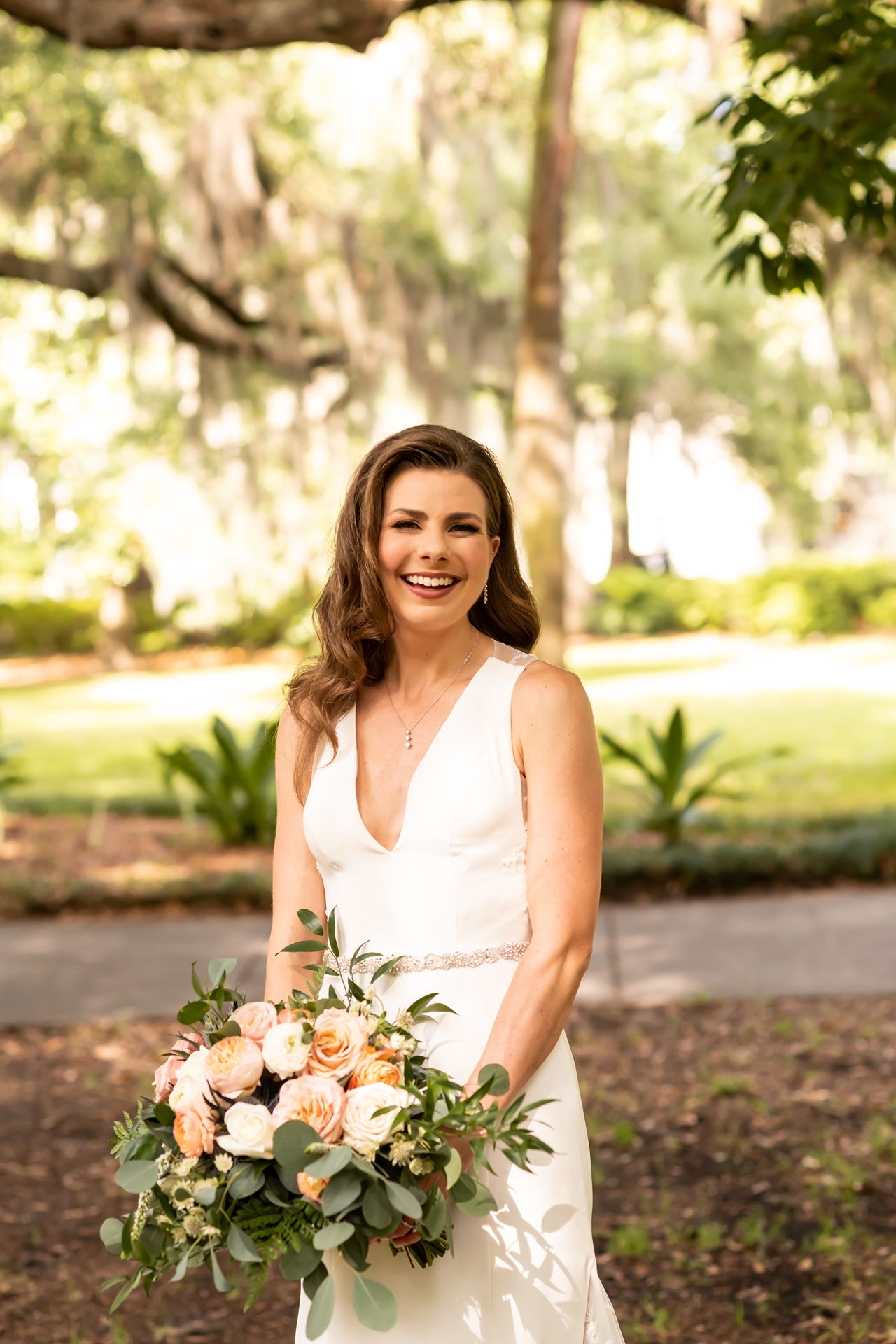 Happy bride wearing white v-neck dress holding peach and white floral bouquet in lush garden setting.