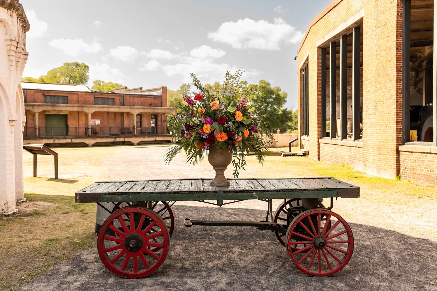 A rustic wooden cart with red wheels displays a large floral arrangement in front of a brick building.