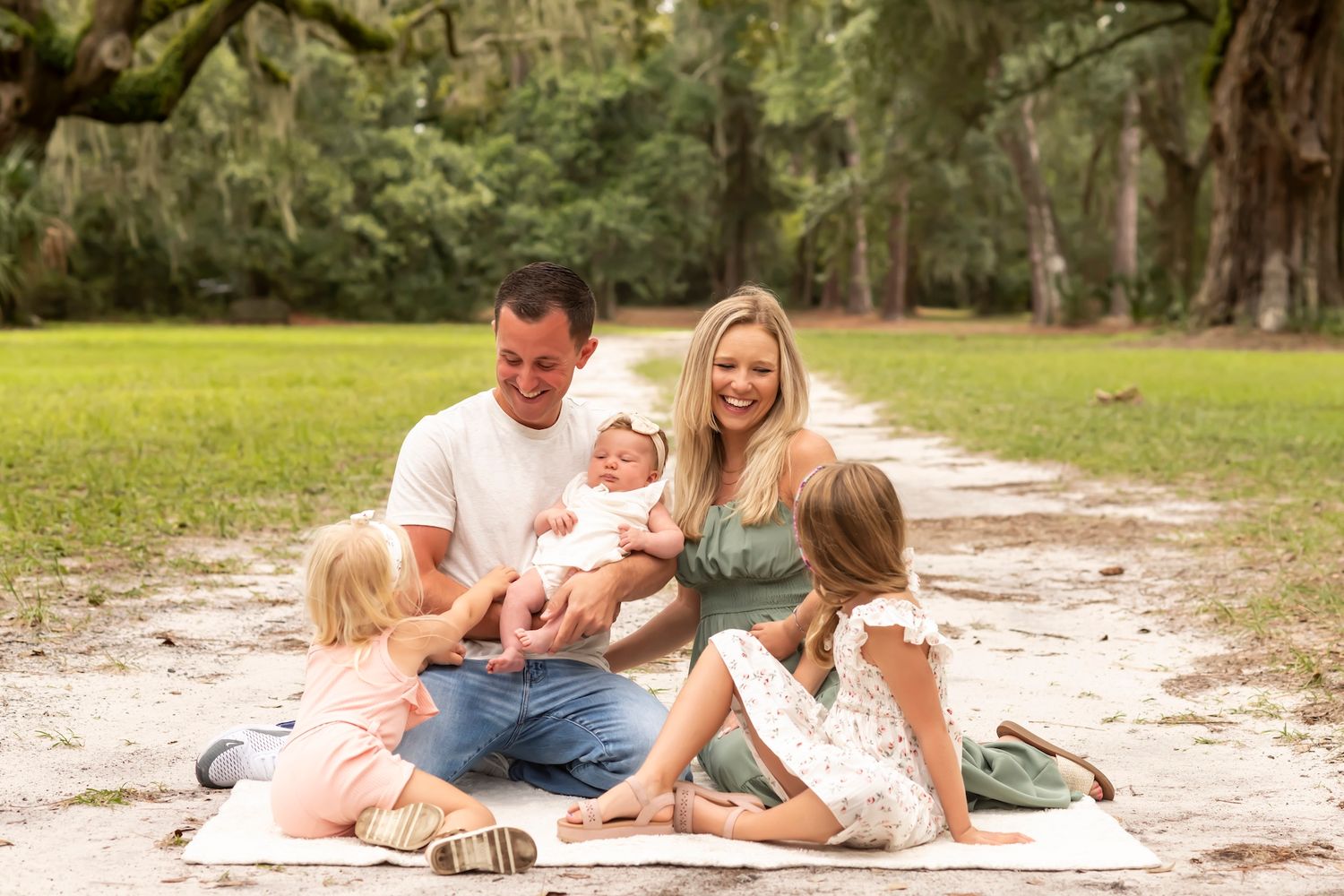 A family sits together on a sandy path surrounded by mossy oak trees and green grass on a sunny day.