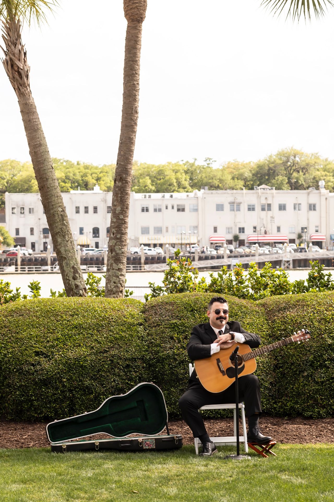 Musician playing acoustic guitar outdoors on lawn with palm trees and white buildings in background.
