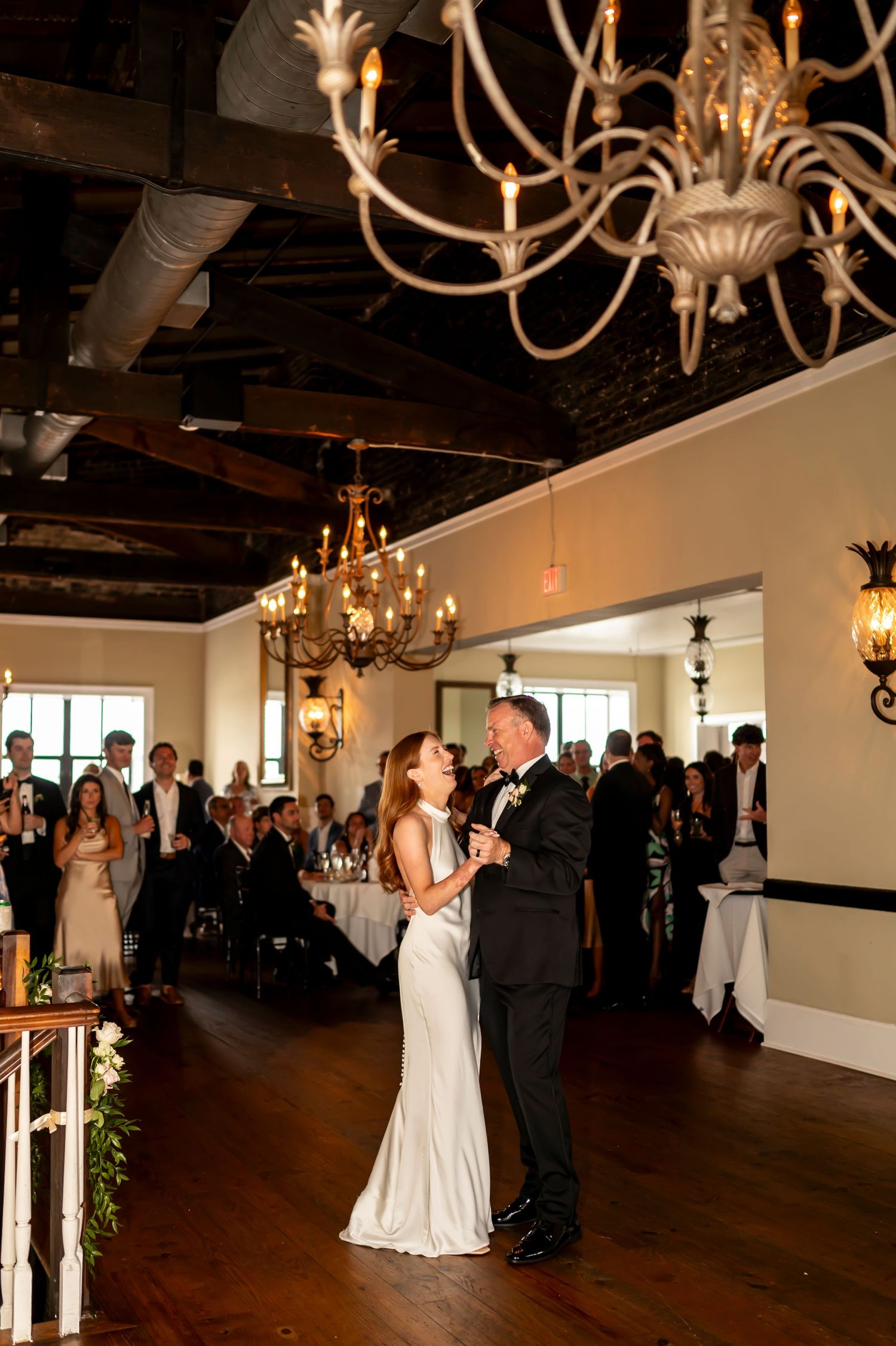 A couple shares their first dance in an elegant reception venue with vintage chandeliers and gathered guests.
