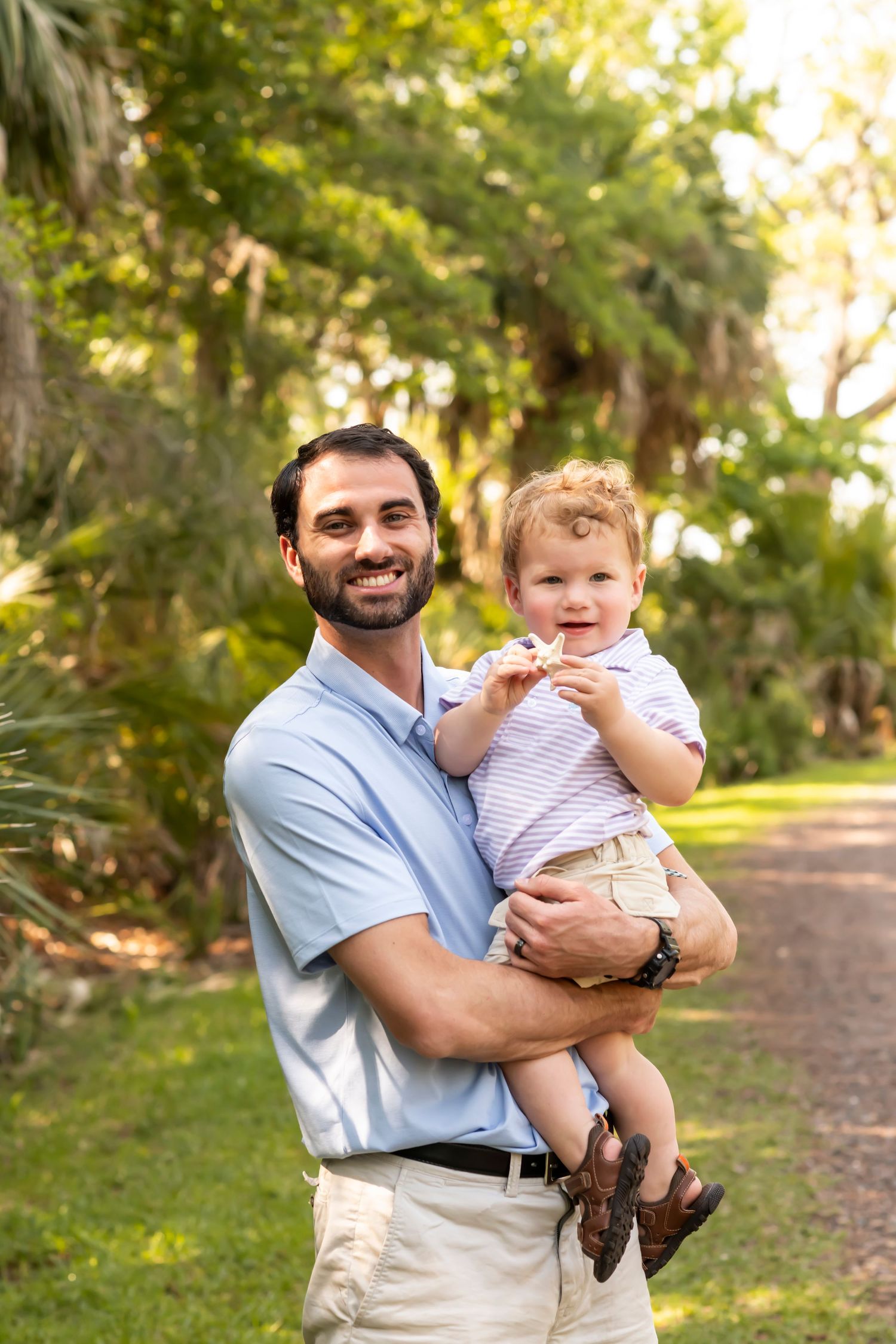 A tender moment captured outdoors on a wooded path as an adult holds a small child in a striped shirt against nature's backdrop.