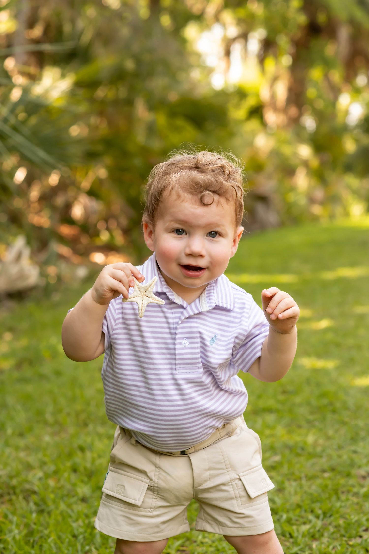 A small child in a striped polo shirt and khaki shorts stands on grass in a sunlit outdoor setting.