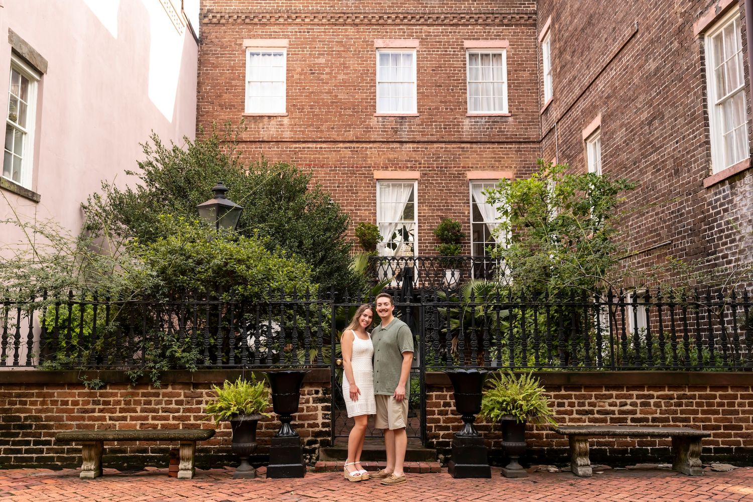 A long row of wooden benches line the brick exterior wall of a historic building next to a manicured garden.