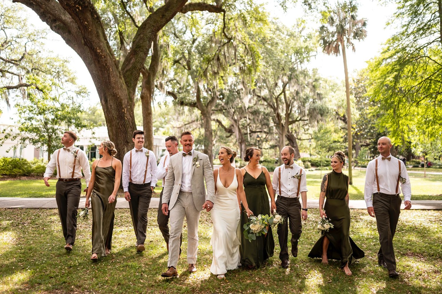 A wedding party wearing black and white attire poses together under Spanish moss-draped oak trees in a sunny park.
