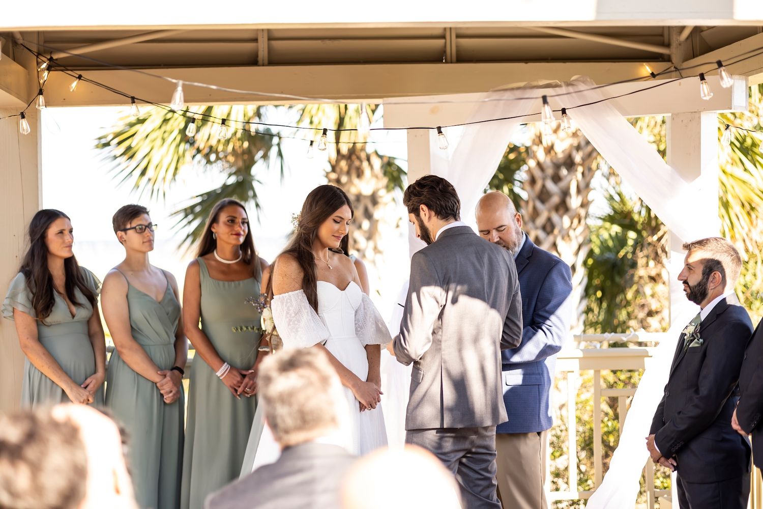 Outdoor wedding ceremony under a covered patio with wedding party in sage green dresses and guests looking on.