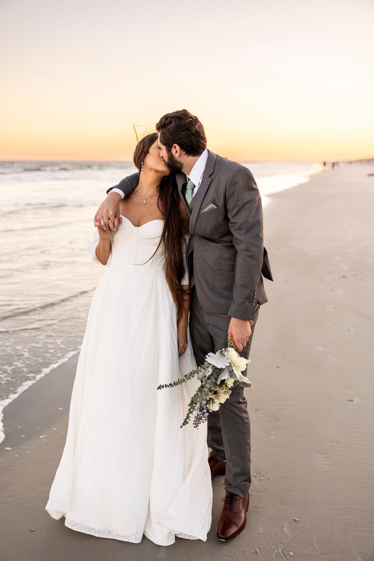 A romantic sunset beach wedding photo with a couple in formal attire holding a white bouquet on the sand.