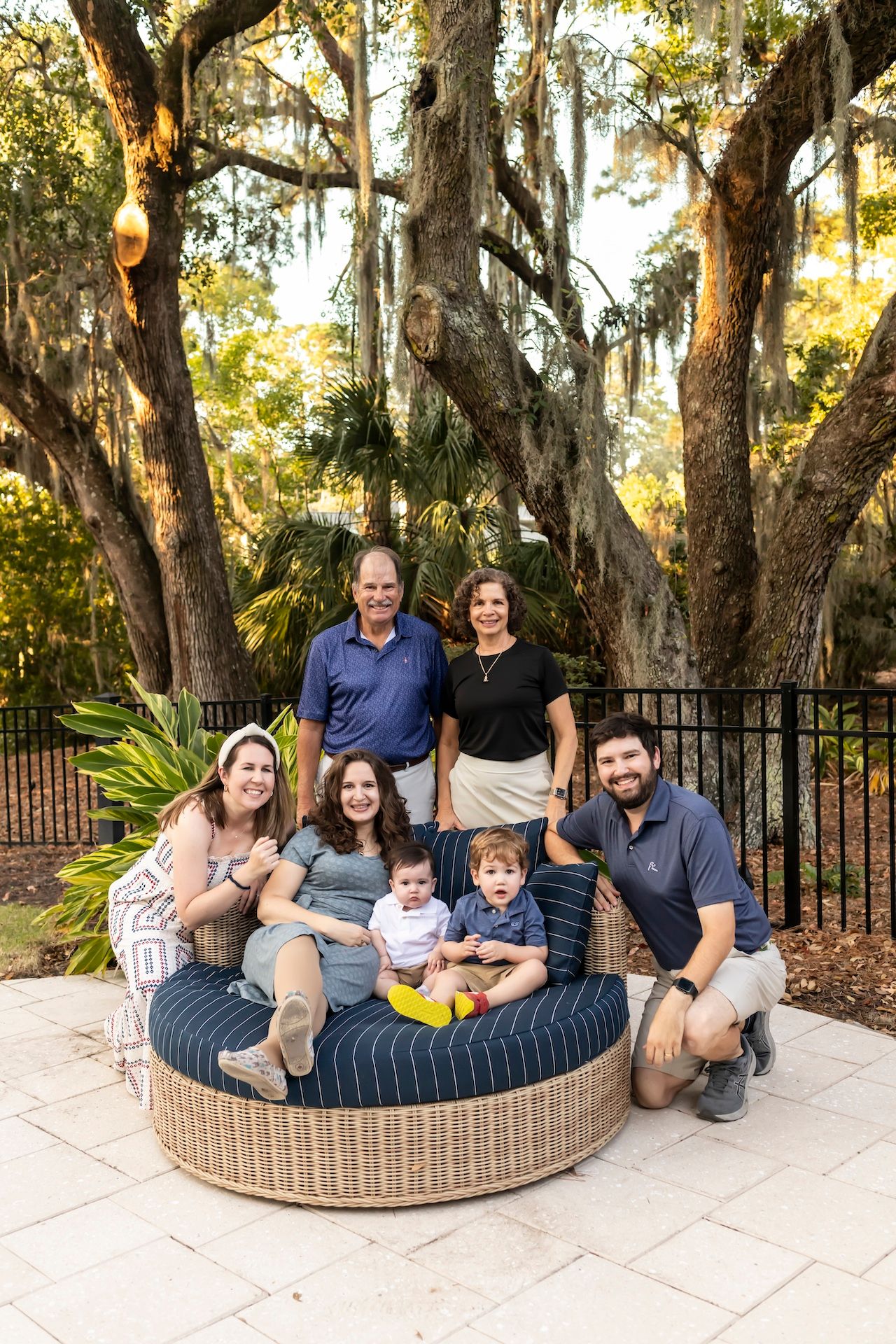A group sitting together in a wicker daybed on a paved patio area surrounded by trees at sunset.