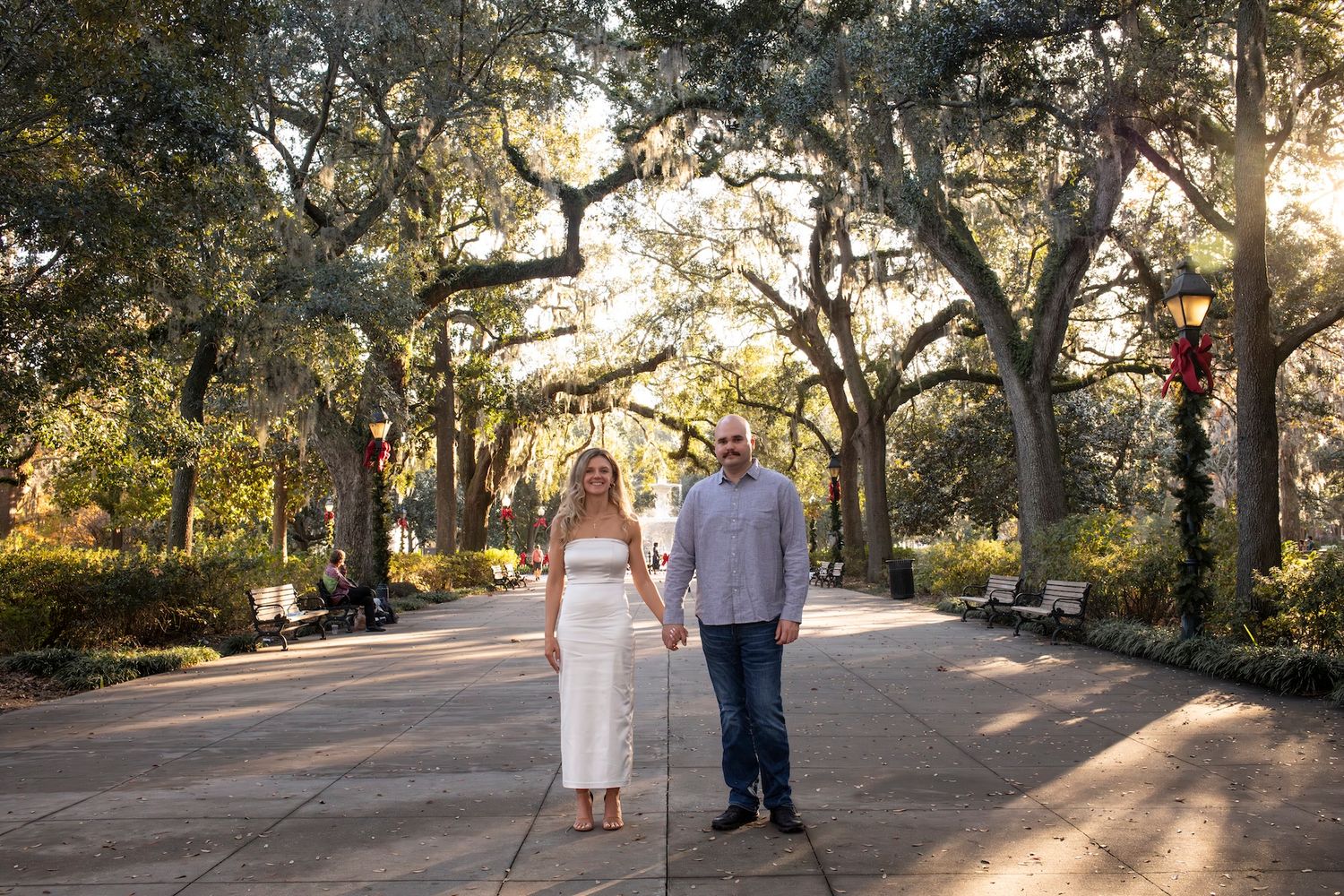 A couple strolls hand in hand down a tree-lined path with sunlight filtering through Spanish moss.