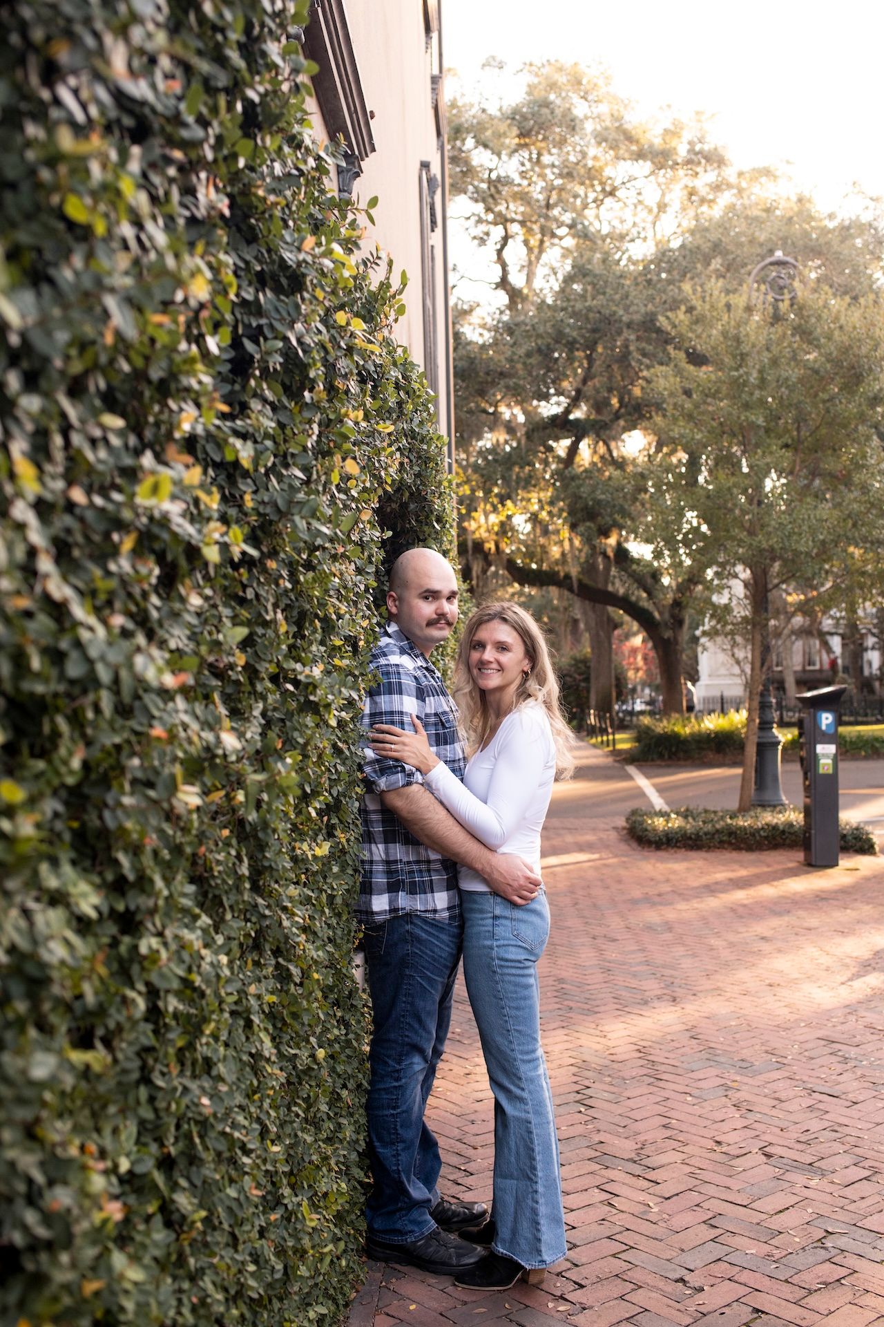 Romantic couple embracing against ivy wall on brick sidewalk during golden hour.