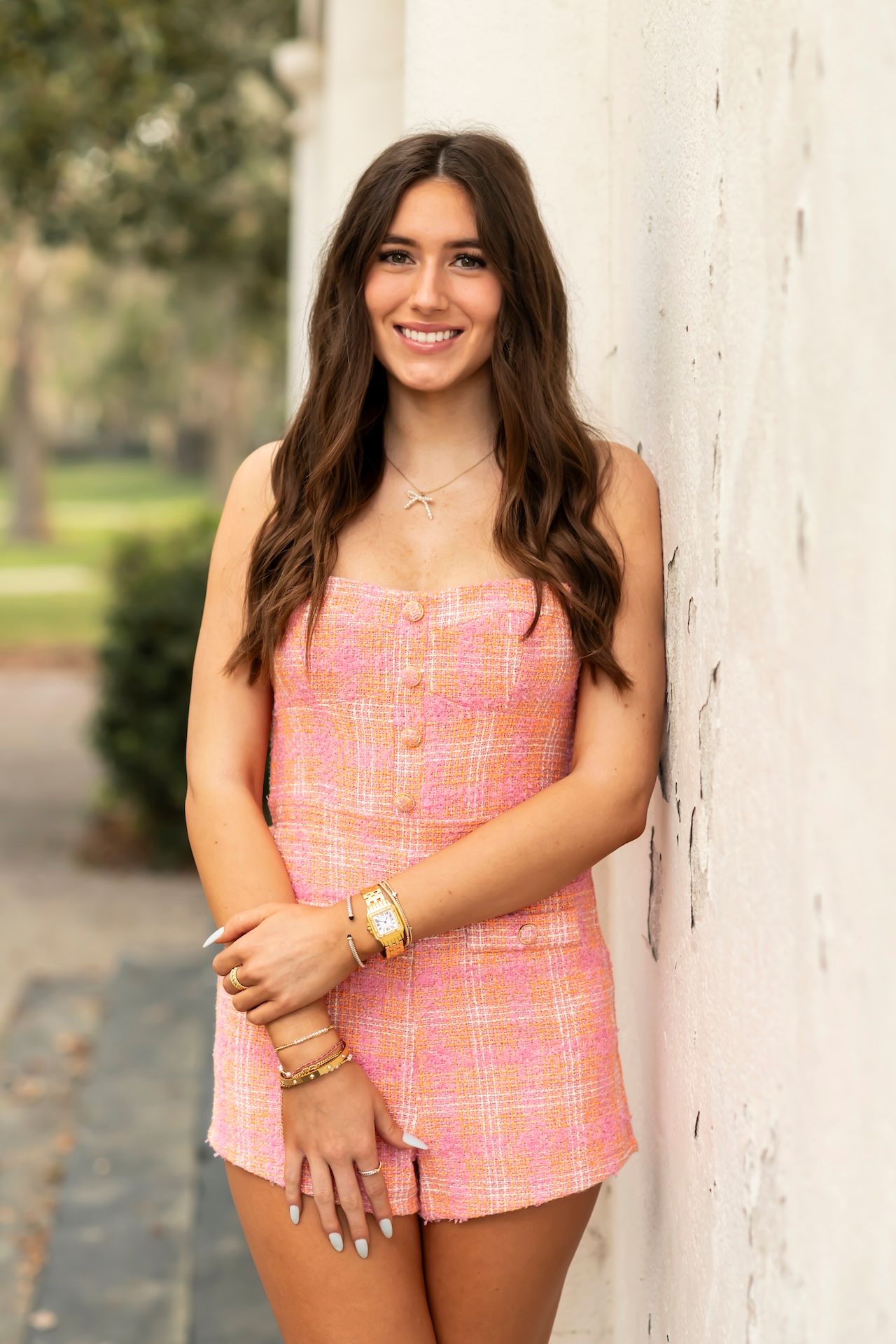 A young woman wearing a pink plaid two-piece outfit smiles while leaning against a white wall outdoors.