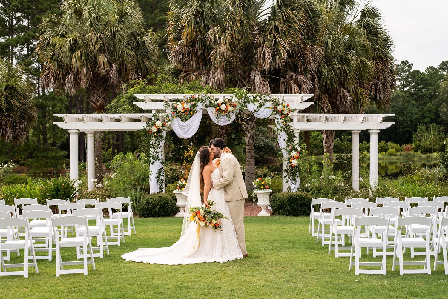 Beautiful outdoor wedding ceremony setup with white pergola arch decorated with flowers and white chairs on a palm tree-lined lawn.