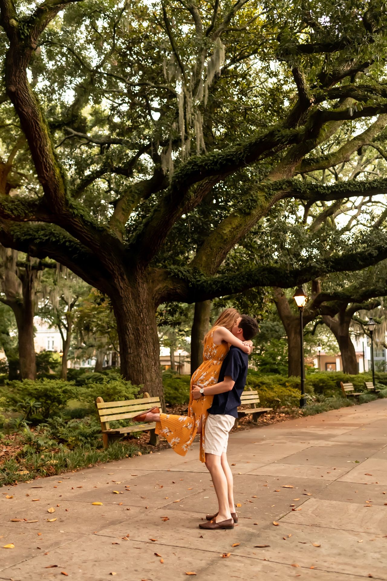 Two figures embrace in a romantic moment on a path beneath sprawling live oak trees in a peaceful park setting.