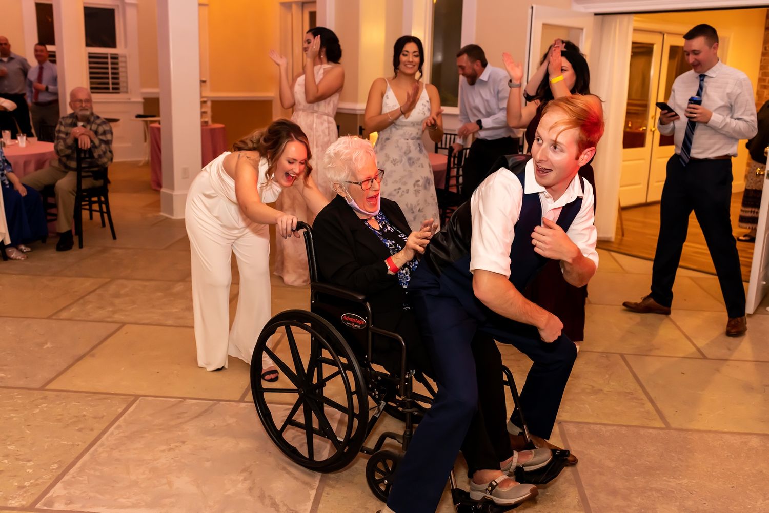 Wedding guests celebrate with elderly person in wheelchair on dance floor during reception celebration.