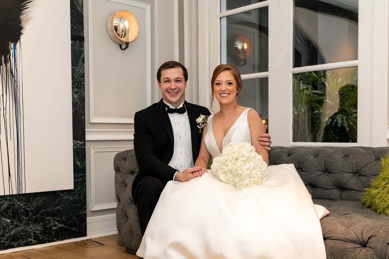 A joyful newlywed couple sits on an elegant gray couch outside a home with white walls and sconce lighting.