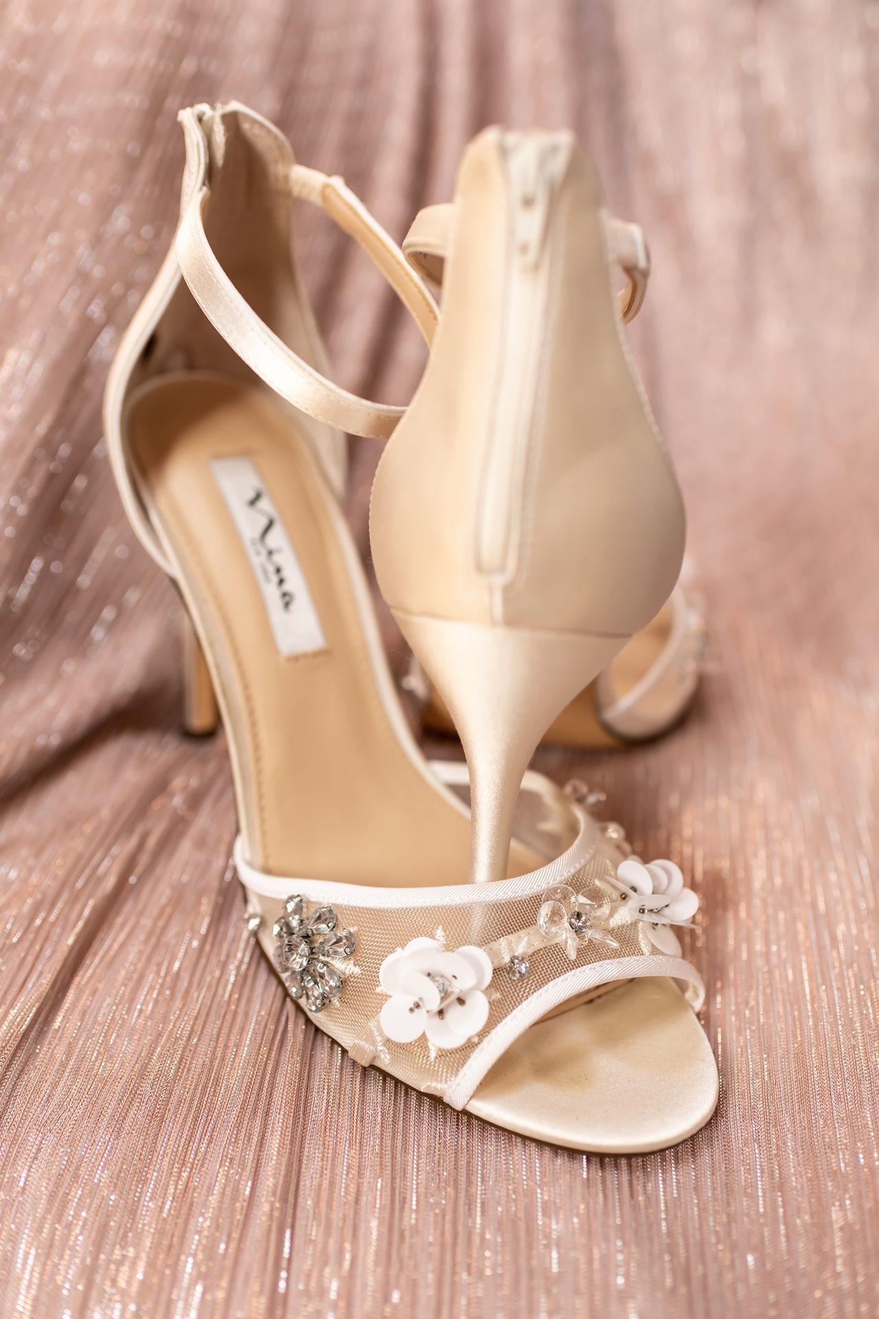 Elegant cream-colored wedding sandals with delicate crystal embellishments placed on a rustic wooden surface.