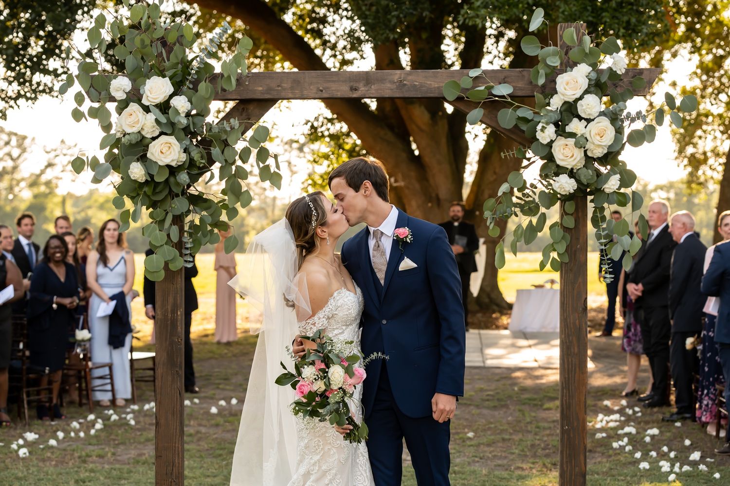 A wedding ceremony under a rustic wooden arch decorated with white roses and florals at sunset.