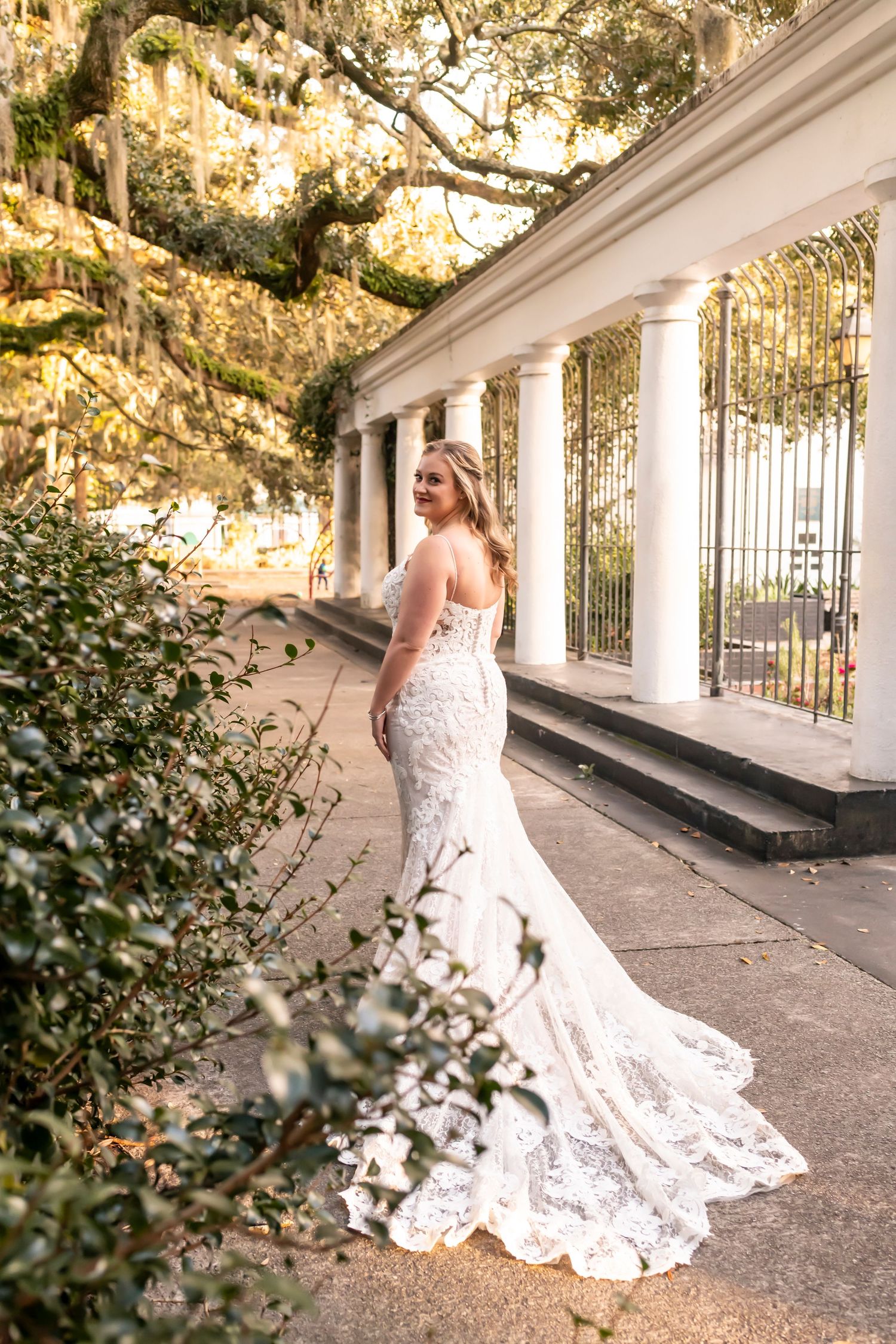 A person in a flowing lace wedding gown poses on a white columned porch with Spanish moss hanging overhead.