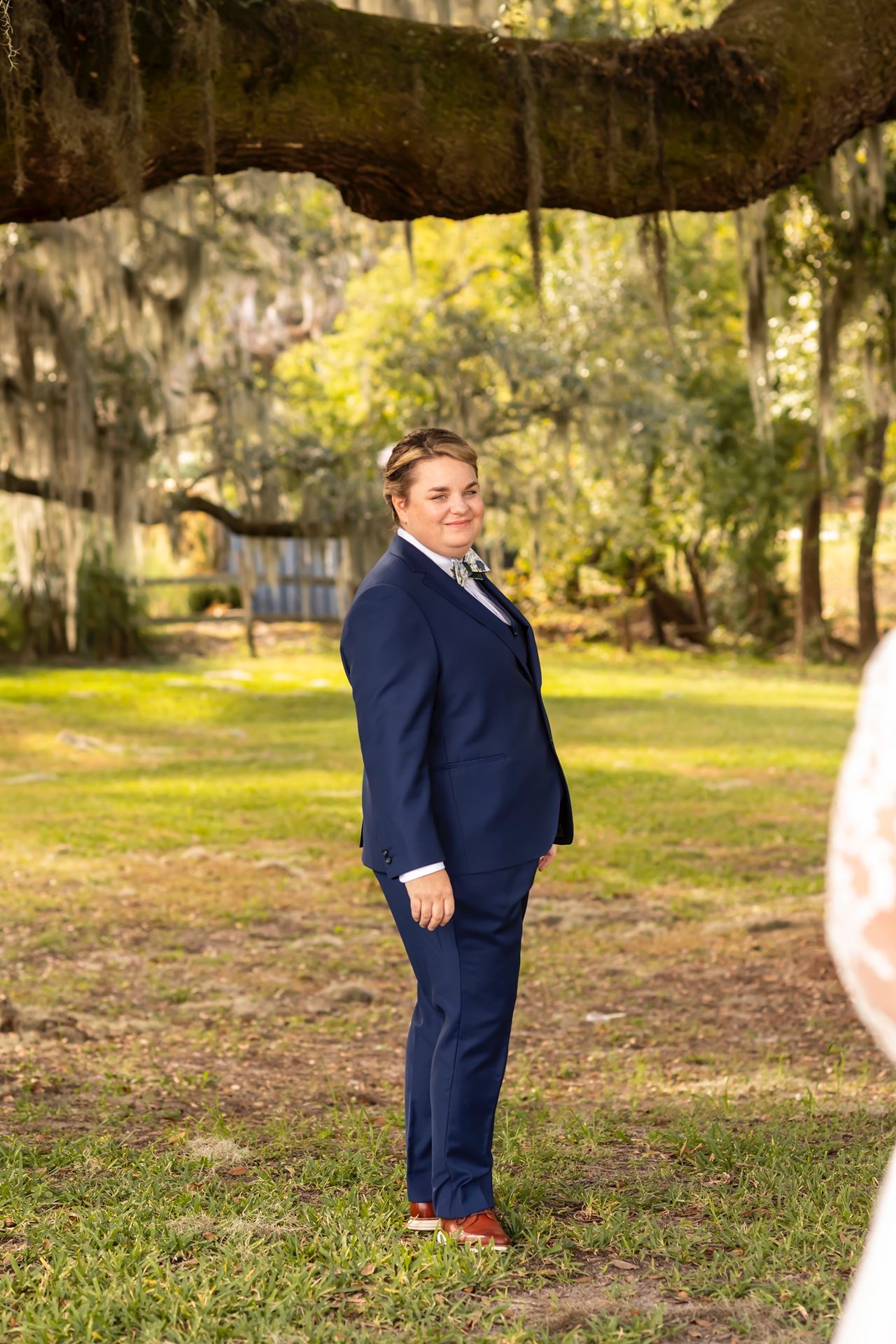 A person in a navy blue suit poses outdoors under moss-draped trees in a sunlit garden setting.