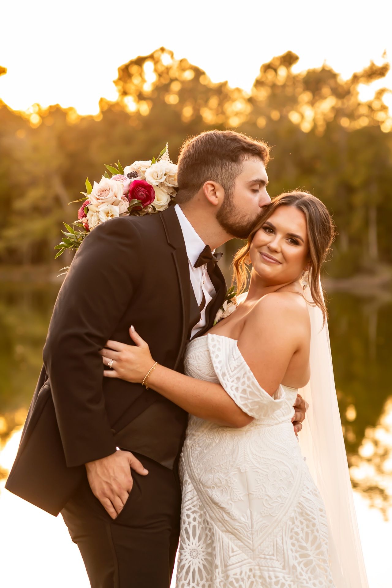 Romantic sunset wedding portrait with couple embracing by a lake while holding a pink and red floral bouquet.