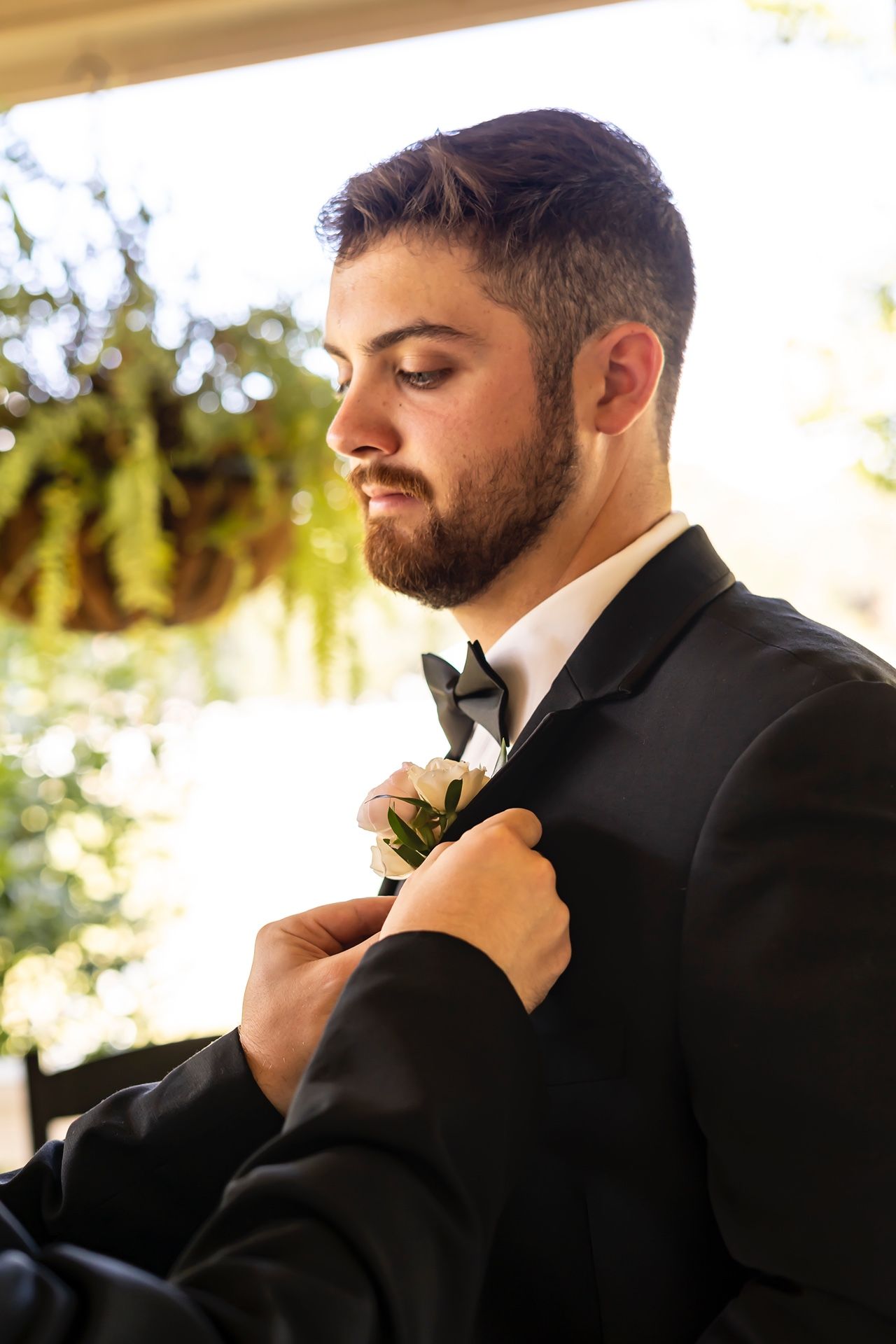 Someone in a black tuxedo and bowtie has a boutonniere being pinned to their lapel in warm outdoor lighting.