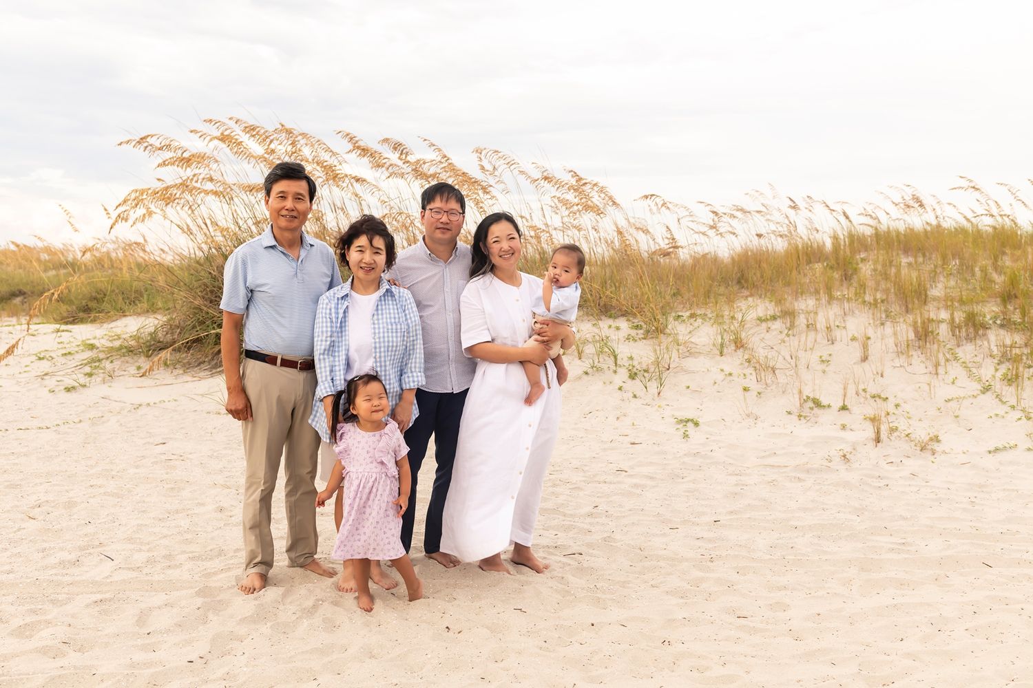 A loving family stands together on a sandy beach with grass-covered dunes in the warm sunset light.