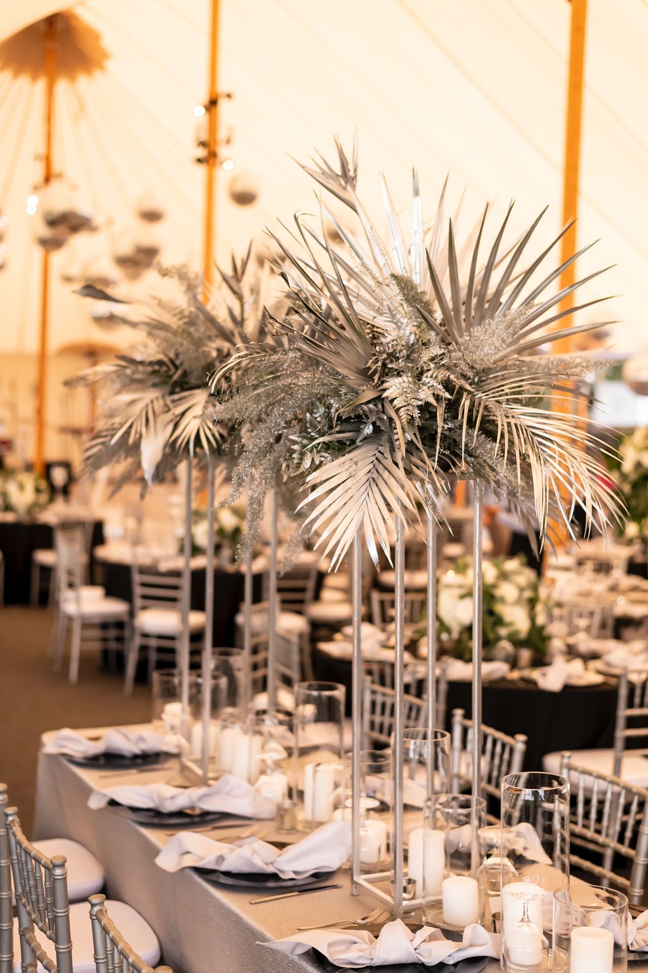 Elegant wedding reception tables decorated with silver palm leaf centerpieces and white chairs in a modern venue.