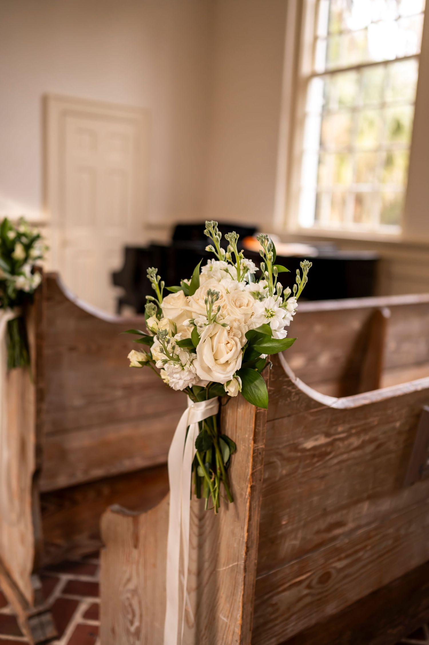 White floral bouquet with ribbons decorates a wooden church pew in a rustic wedding ceremony setting.