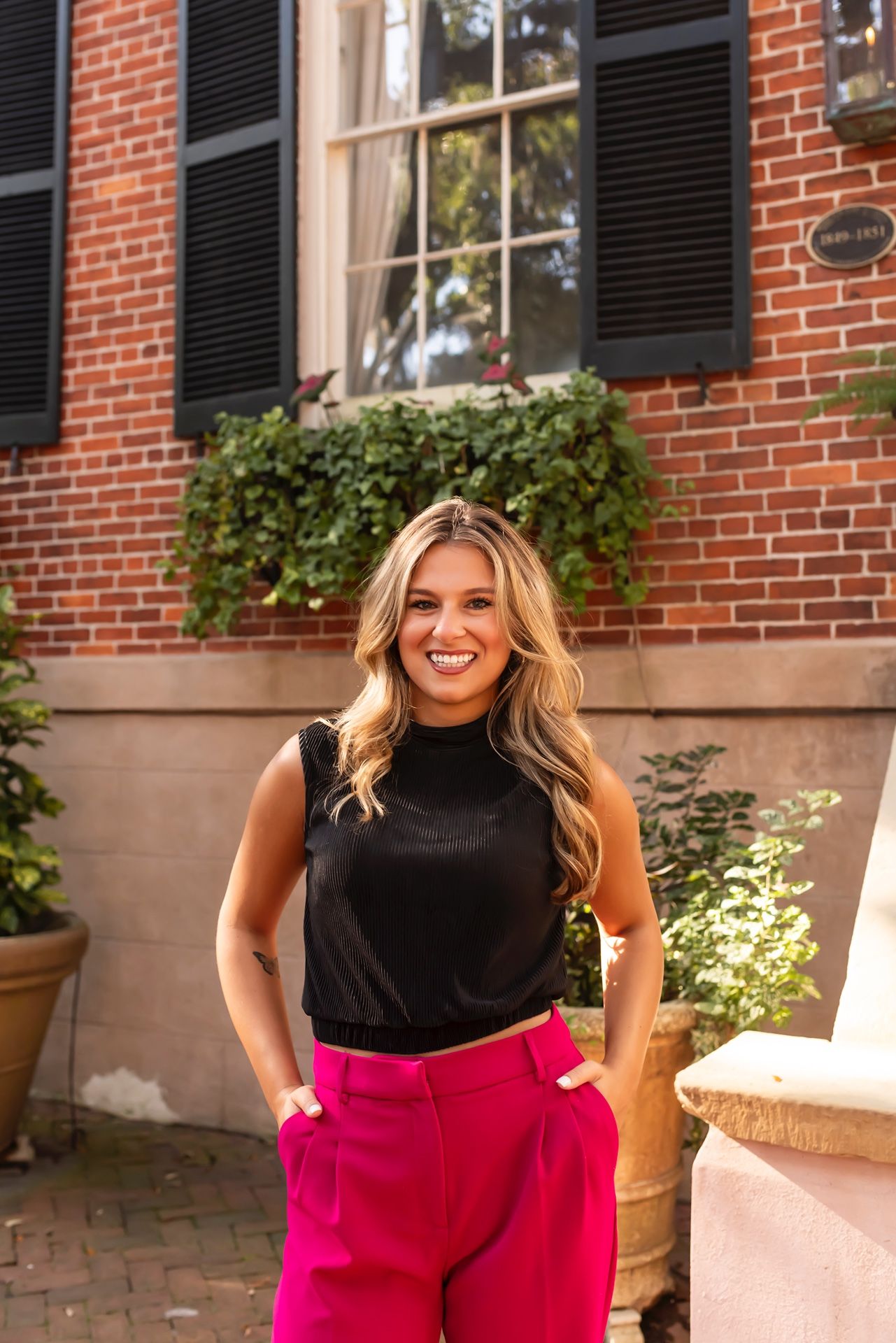 A woman in a black sleeveless top and pink pants poses in front of a brick building with black shutters and window boxes.