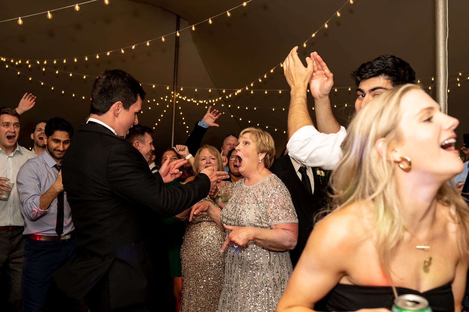Wedding guests dancing and celebrating under romantic string lights at an indoor reception venue.