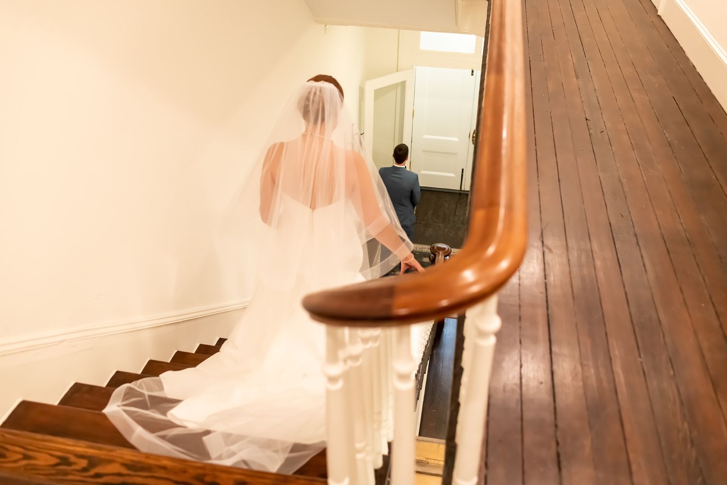 A bride in a flowing white dress descends a wooden staircase with curved banister in an elegant indoor setting.