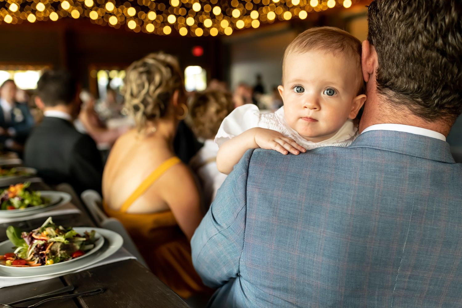 A small child sits on someone's shoulder at a restaurant with glowing lights and plates of food visible in background.