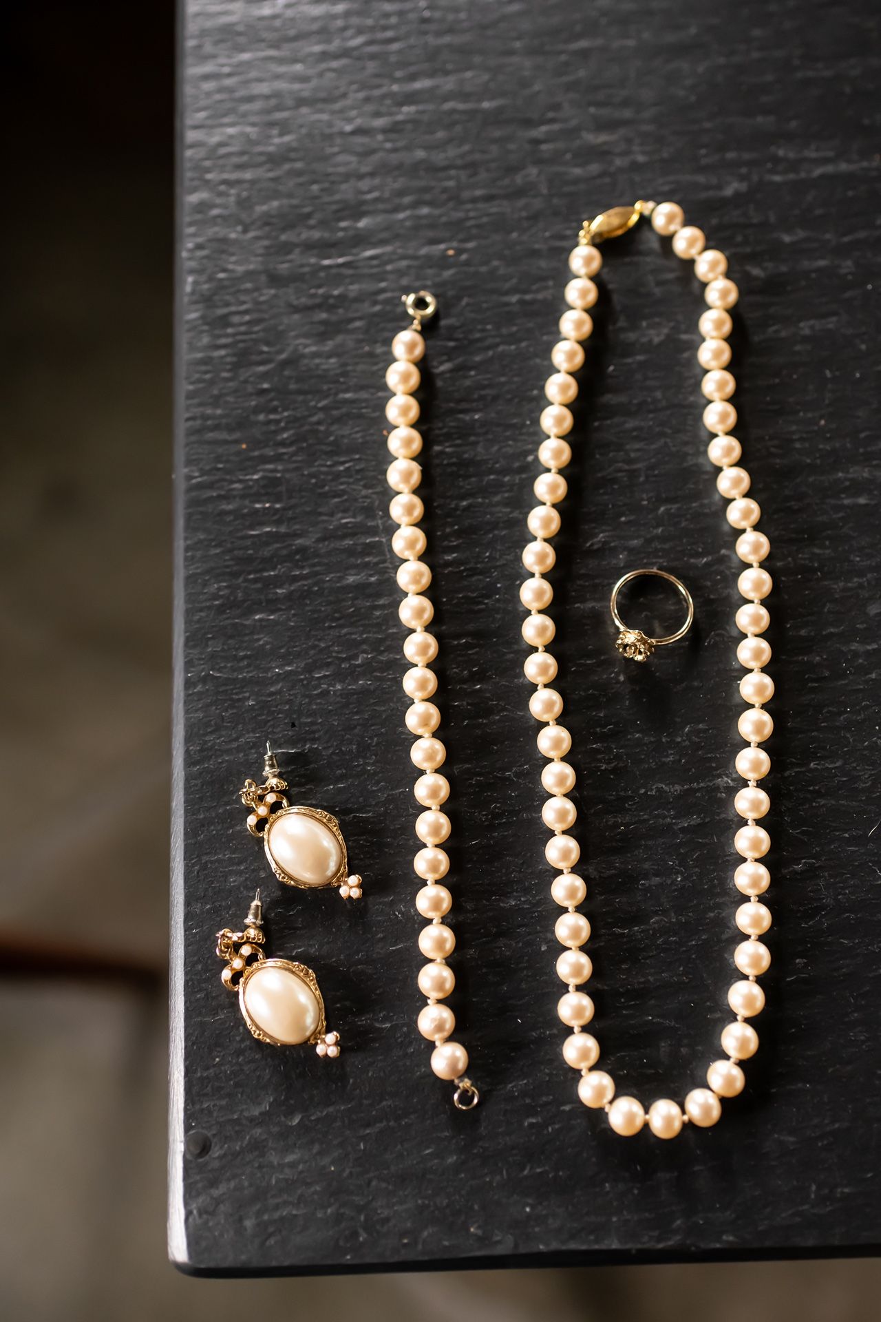 A pearl necklace and matching pearl earrings with a gold ring displayed on a dark slate surface.