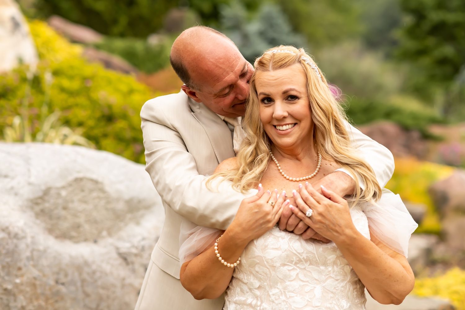 A couple embraces outdoors in wedding attire amid a sunny garden setting with yellow flowers and stone features.