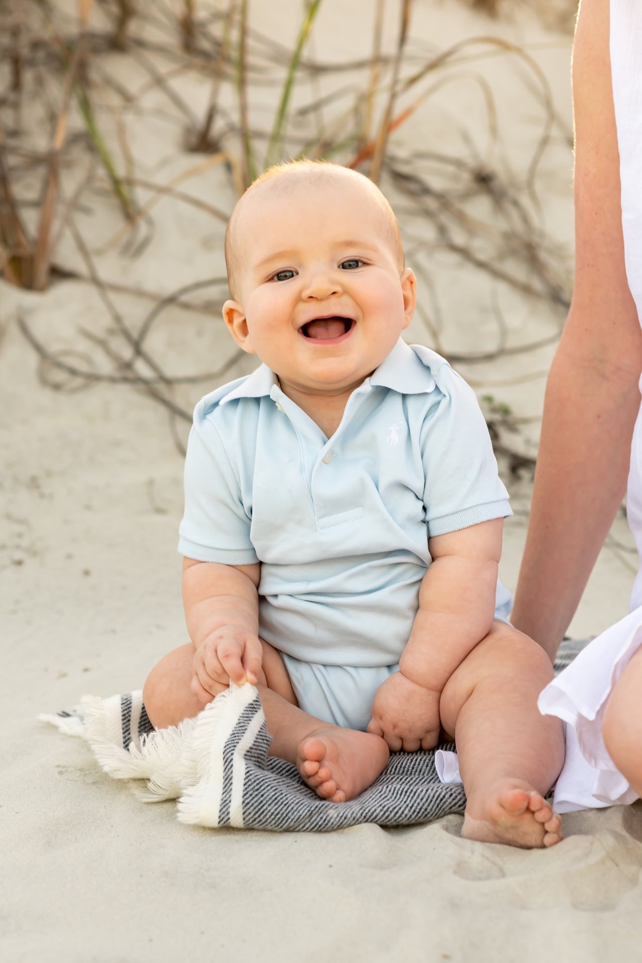 Laughing baby wearing light blue polo shirt sitting on sandy beach with adult beside them.
