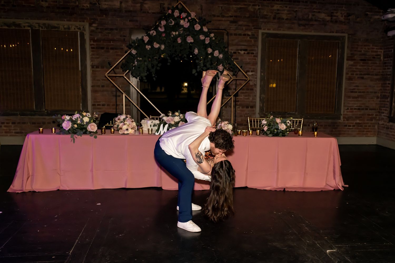 A couple dances at their wedding reception in front of a pink-draped table decorated with floral arrangements.