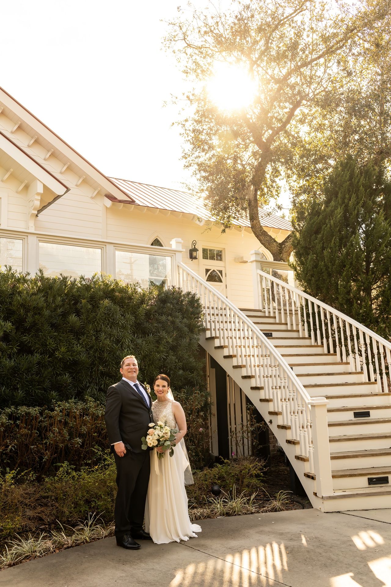 A couple shares a romantic moment beside white exterior stairs during a golden sunset at an elegant venue.