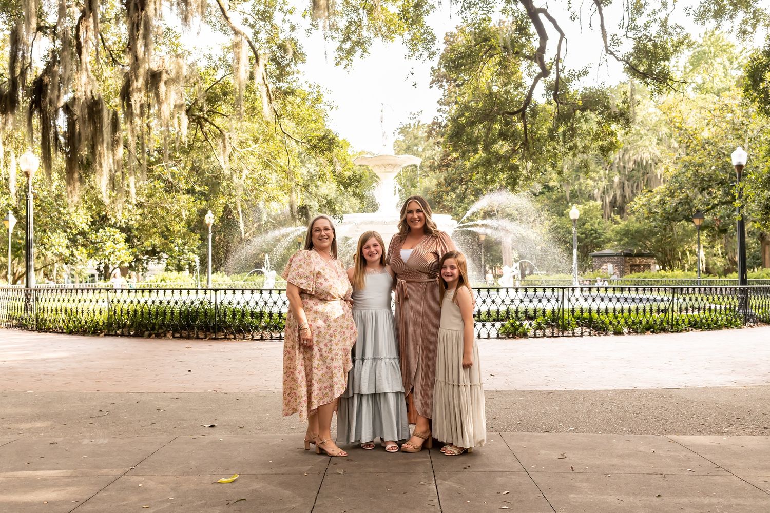 Three people in elegant dresses stand together at an outdoor venue with Spanish moss draped trees and white fence.