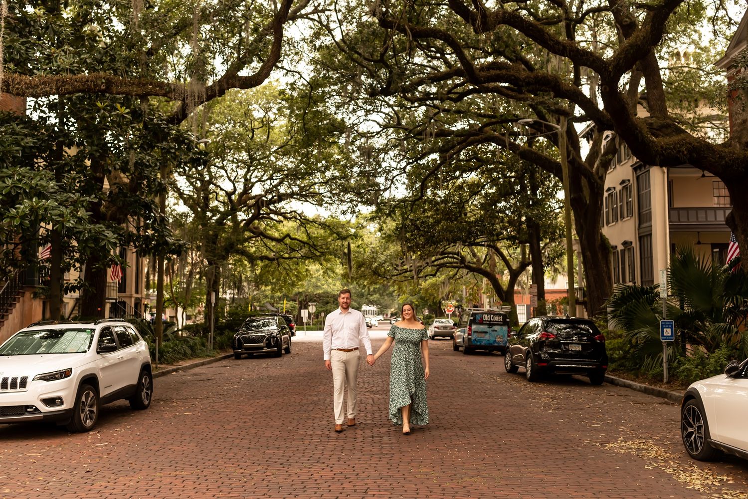 People stroll down a charming tree-lined brick street in Savannah Georgia with parked cars and historic homes.