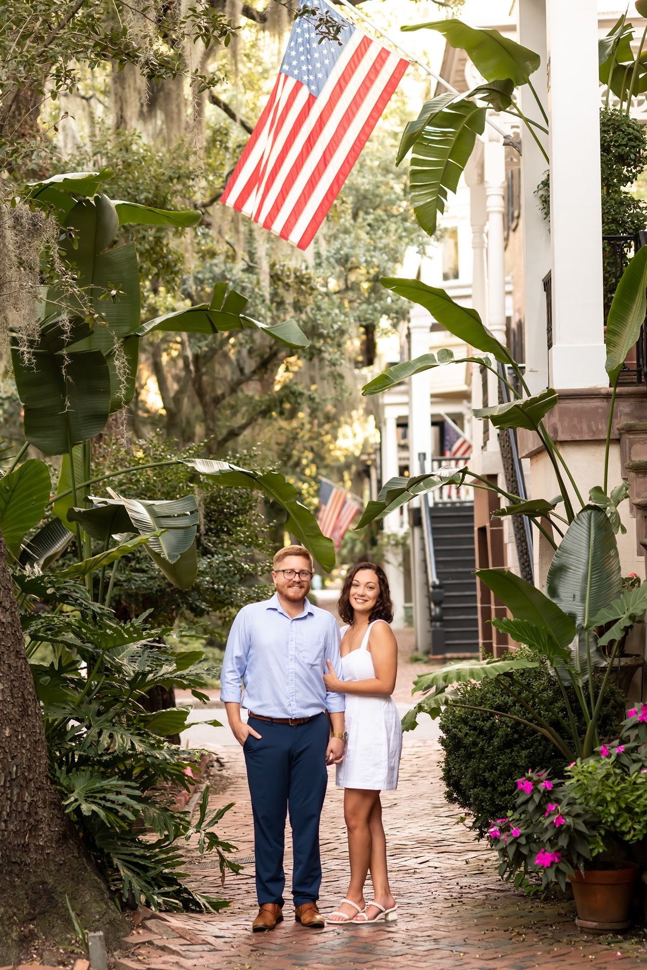 A couple stands on a charming garden pathway with lush tropical plants and an American flag in the background.