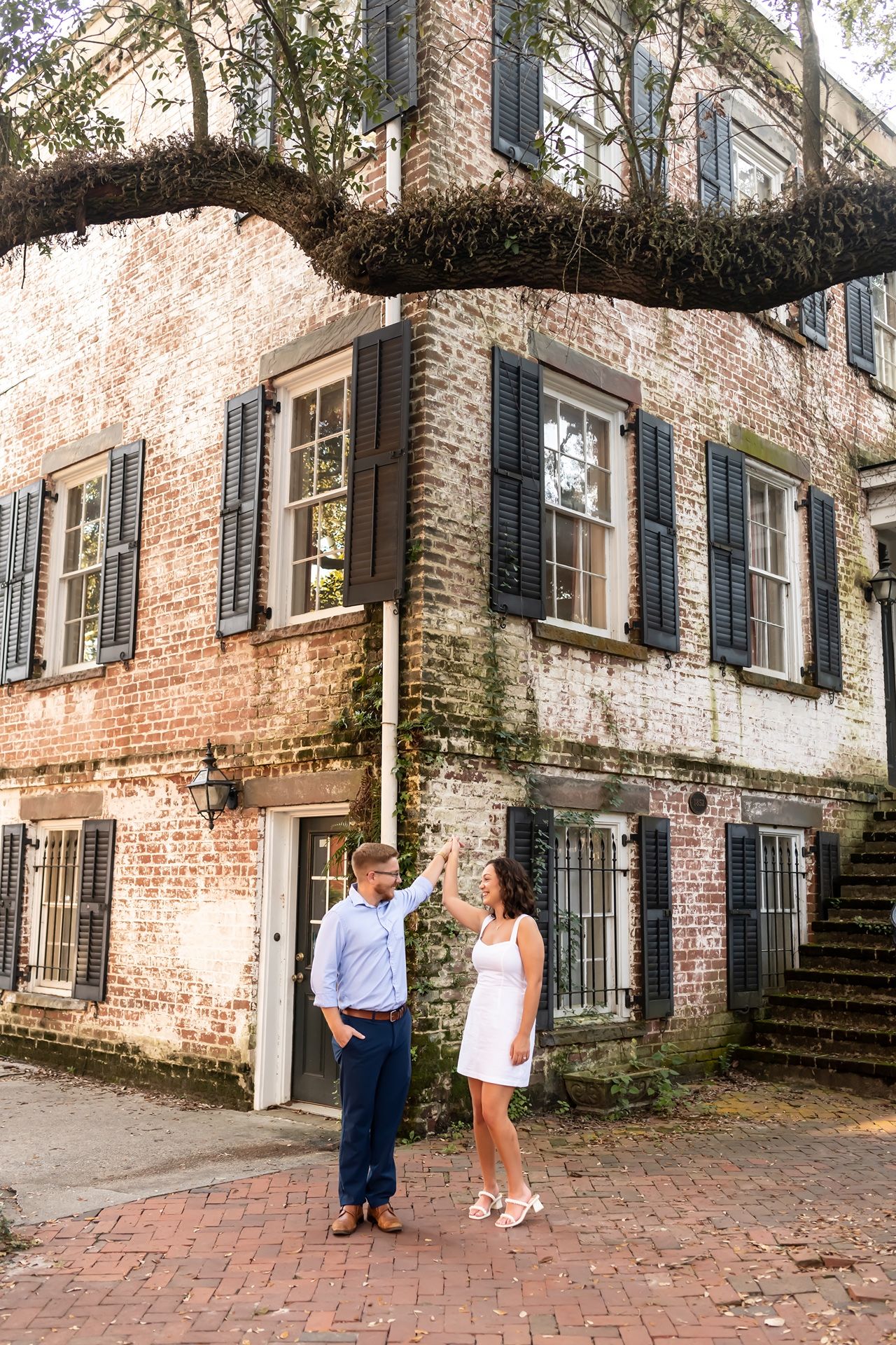 Two people stand on a brick sidewalk beside a historic Charleston building with moss-covered limbs and black shuttered windows.