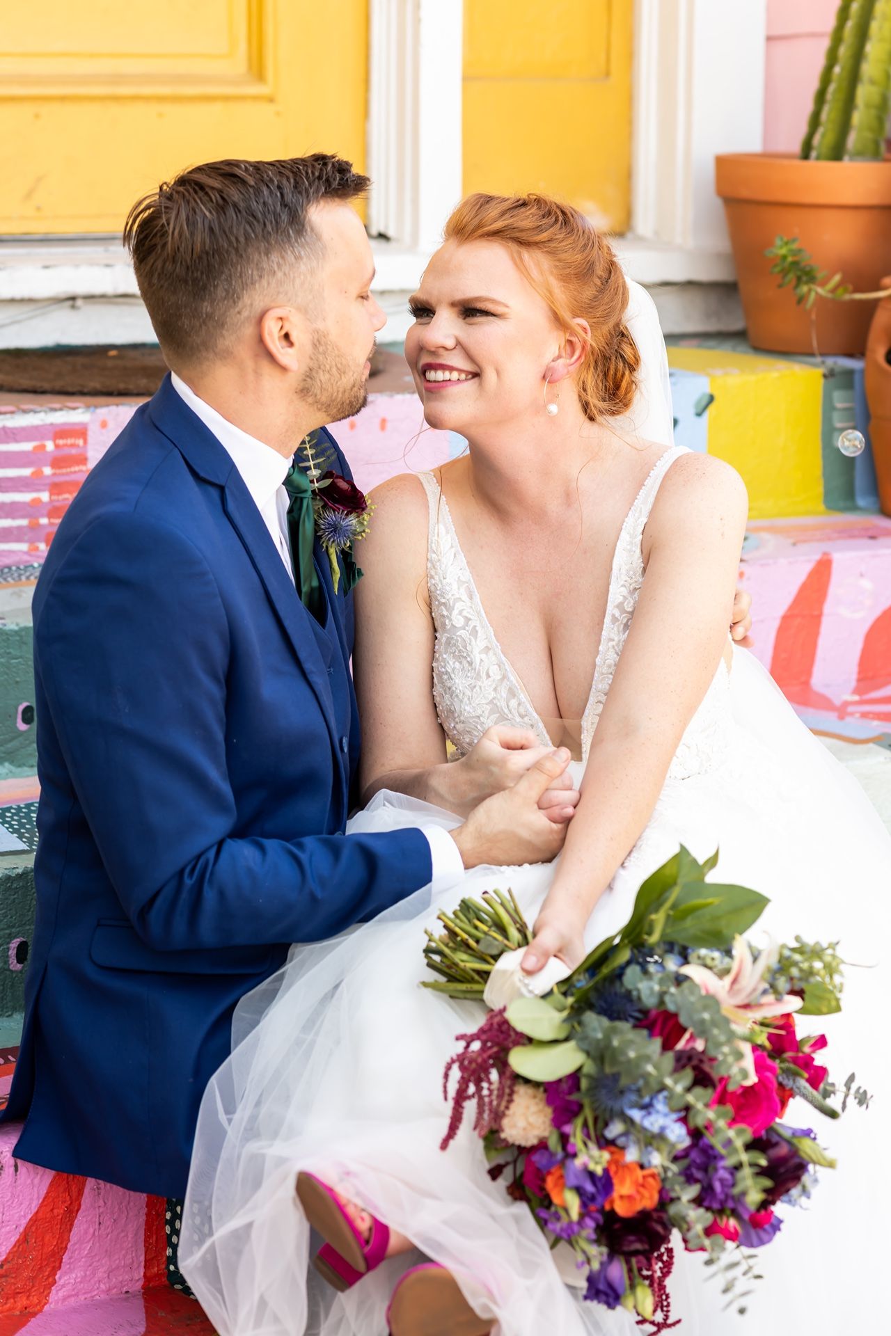 A wedding couple shares an intimate moment outdoors against a bright yellow wall with colorful floral bouquets.