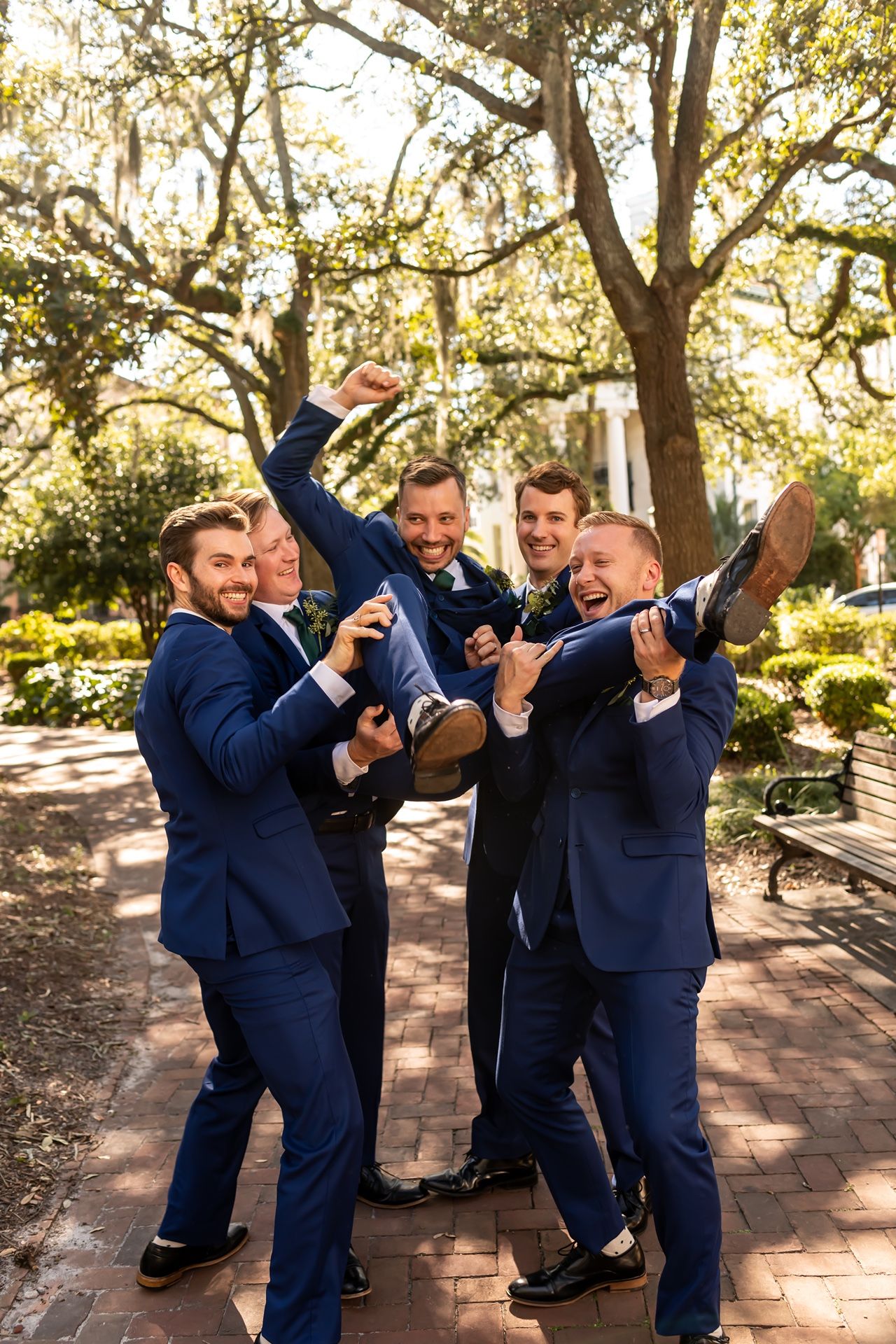 A group in matching navy blue suits strike playful poses outdoors under moss-draped trees on a brick pathway.