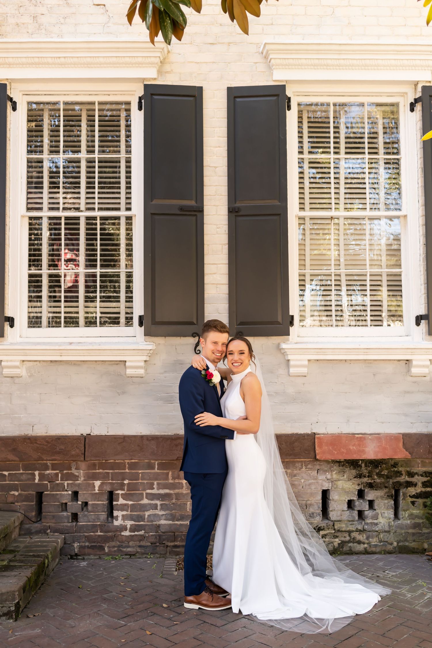 A lovely couple embraces against a white colonial-style house with black shutters and exposed brick foundation.