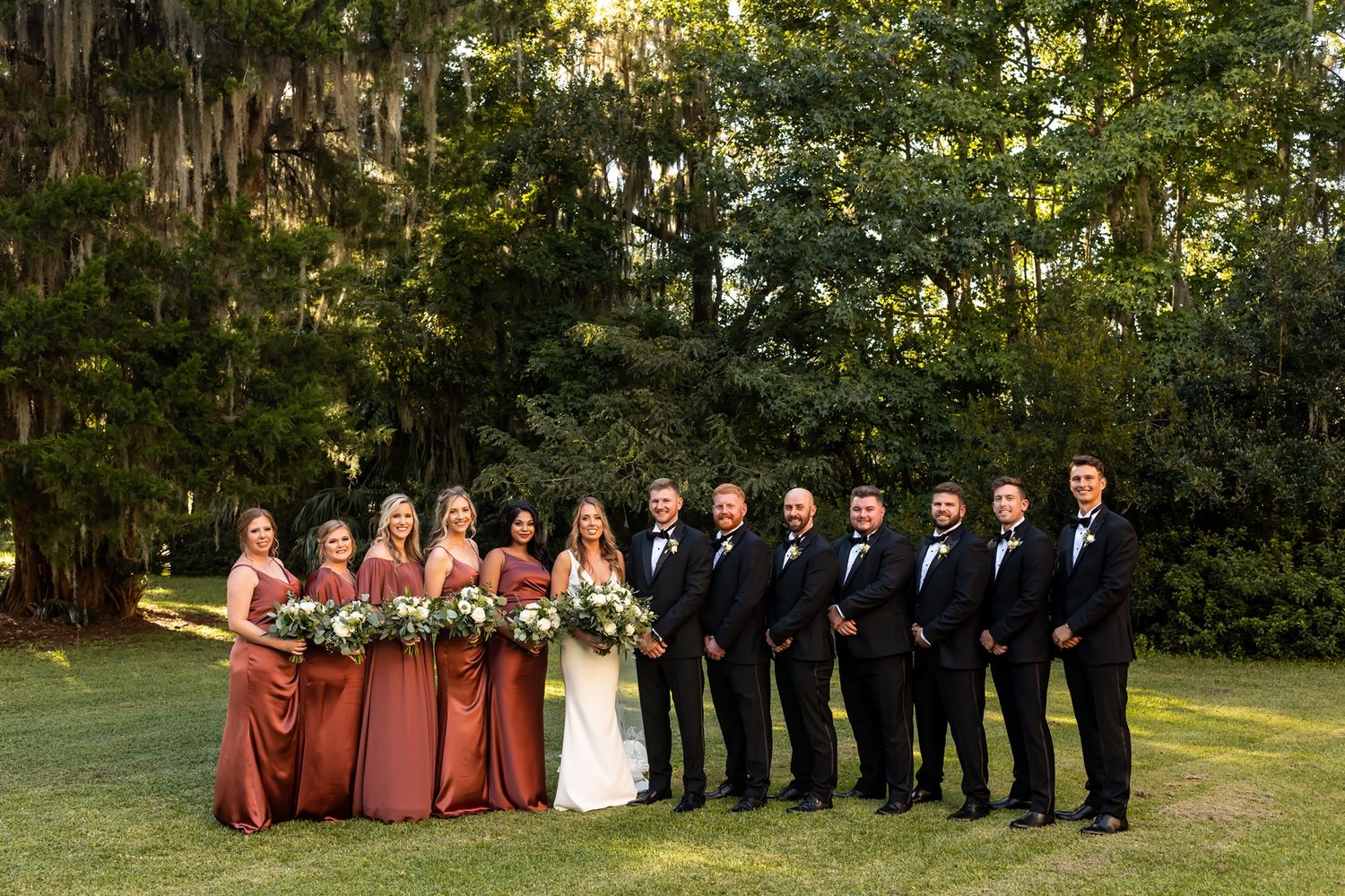 Wedding party in rust-colored dresses and navy suits poses together on a scenic lawn with mossy trees in background.