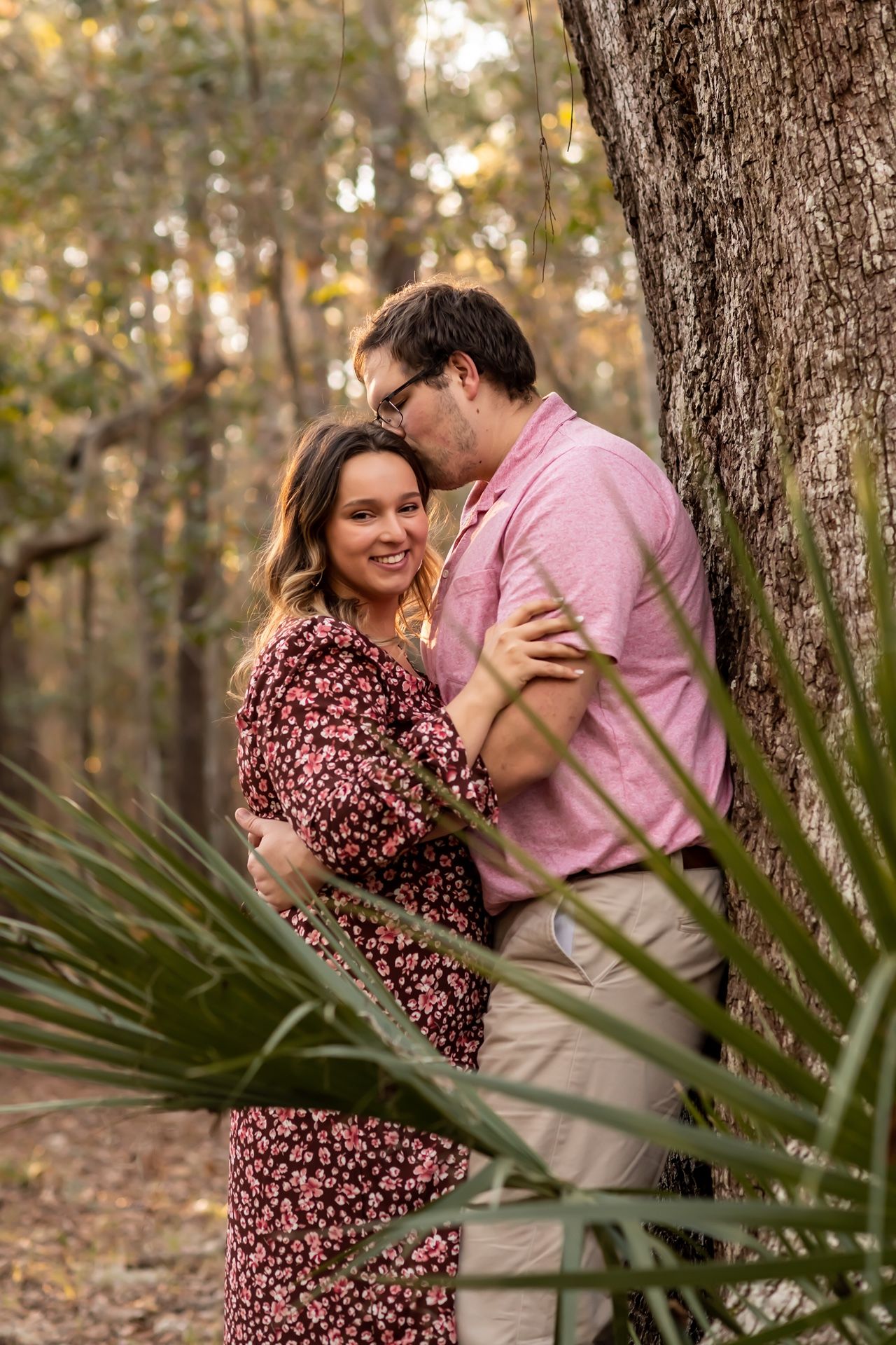 A couple embraces in front of a large tree during an outdoor photo session with palm fronds in the foreground.