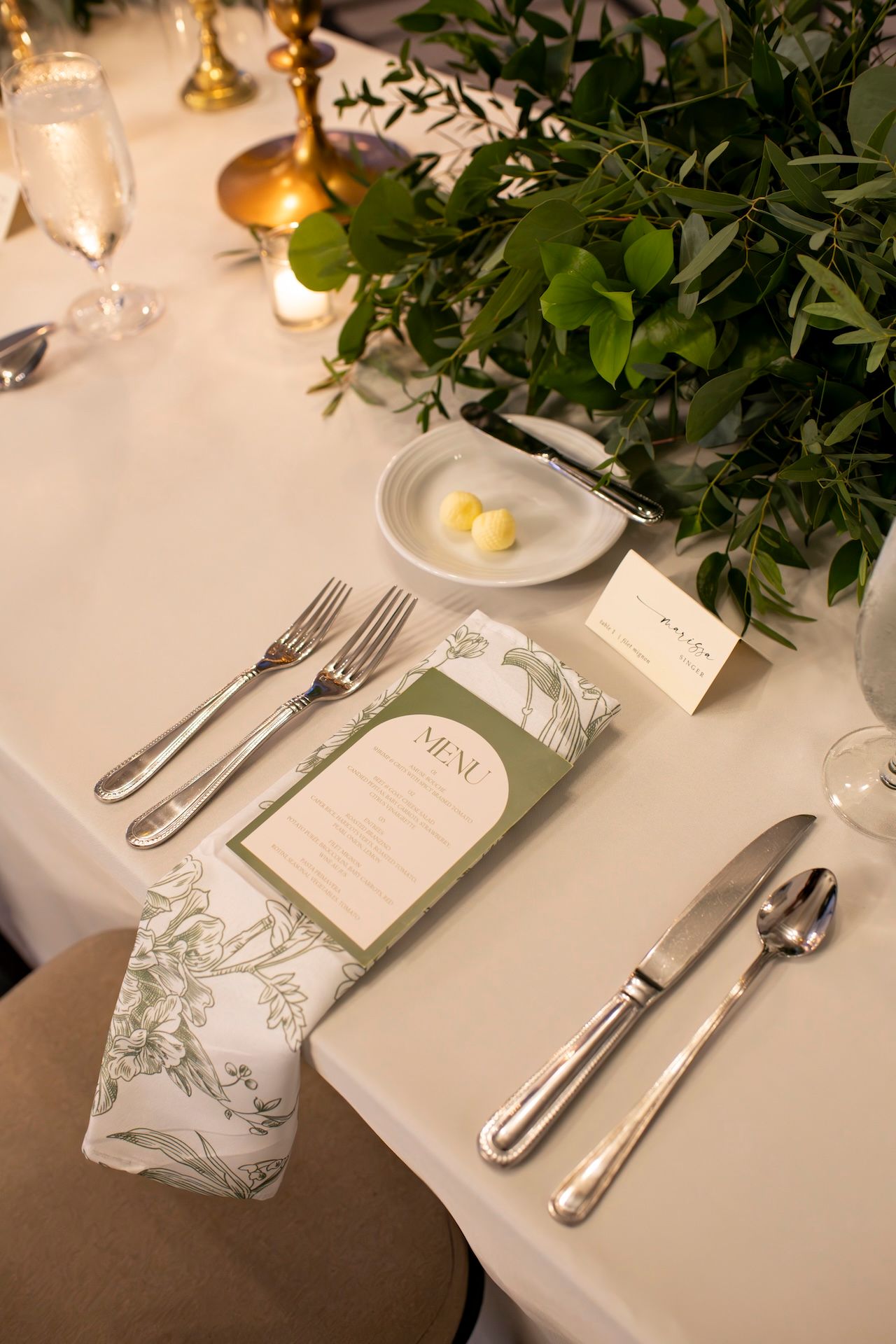Close-up of formal place setting with white menu card, silverware, and greenery on white tablecloth.