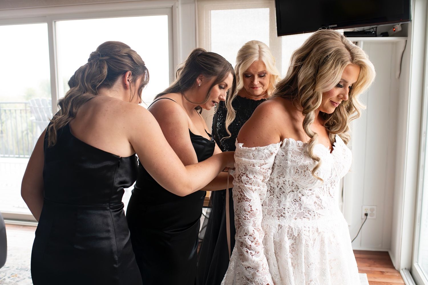 Bridesmaids in black dresses help bride into her white wedding gown near bright windows.